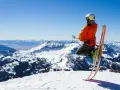 Un skieur en l'air, prêt à dévaler la pente. Il porte un casque et une veste orange vif, rappelant les règles pour skier en toute sécurité. Le paysage montagneux enneigé est spectaculaire.