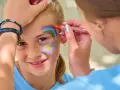 Une petite fille souriante se fait maquiller sur la joue un arc-en-ciel et des fleurs, au stand maquillage d'une kermesse.