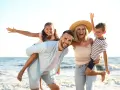 Famille souriante sur la plage, père portant sa fille sur le dos, mère tenant son fils.