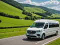 Minibus blanc sur une route de campagne verdoyante, avec des collines en arrière-plan.