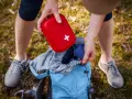 Une personne prépare son sac à dos pour une randonnée, rangeant une trousse à pharmacie enfant rouge avec une croix blanche.