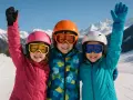 Trois enfants souriants, équipés pour le ski, célèbrent leurs vacances scolaires en Autriche. Leurs casques et lunettes colorés brillent sous le soleil.
