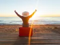 Personne assise sur une valise rouge sur une plage au coucher du soleil, bras levés en signe de victoire.