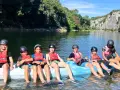 Des enfants en gilets de sauvetage et casques sur un raft bleu sur une rivière, entourés de verdure et de falaises.