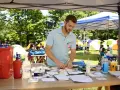 Un homme en polo bleu prépare du matériel médical sur une table, avec des tentes et des enfants en arrière-plan.