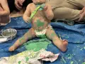 Bébé couvert de peinture verte, tenant un pinceau, assis sur une bâche bleue.