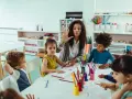 Une animatrice explique une activité créative à des enfants autour d'une table. Son rôle est d'éveiller leur curiosité.