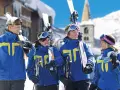Quatre jeunes skieurs souriants en tenue bleue et jaune, portant des skis et des bâtons, dans une station de ski enneigée.