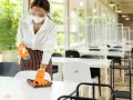 Employée portant masque et visière nettoie une table, respectant la réglementation hygiène cantine scolaire. Des séparateurs sont installés.