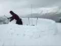Person construisant un igloo en blocs de neige, avec des bâtons de ski plantés.