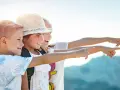Trois enfants souriants, portant des sacs à dos, pointent du doigt vers la droite, sous un ciel ensoleillé.