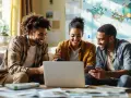 Trois jeunes adultes souriants regardent un ordinateur portable sur un canapé, une carte du monde au mur.
