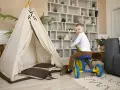 Un petit garçon sur un tricycle bleu et jaune s'amuse dans un espace de jeux, près d'une tente tipi et d'un canapé. Ce centre de vacances et de loisirs est idéal pour les enfants.