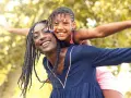 Une femme porte une enfant sur son dos, toutes deux souriantes, avec des arbres en arrière-plan.