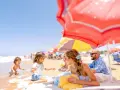 Famille souriante sur une plage ensoleillée, sous des parasols colorés, avec des enfants jouant dans l'eau.