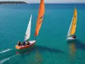 Deux voiliers naviguent sur une mer turquoise, l'un avec une voile orange, l'autre jaune.
