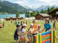 Animation colonie de vacances : des enfants rient en jouant avec un ballon, une monitrice les supervise. Paysage de montagne et lac en arrière-plan.