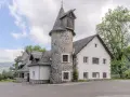 Bâtiment blanc avec une tour ronde en pierre et un toit en ardoise, entouré d'arbres et de verdure.