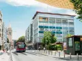 Un bus rouge circule dans une rue bordée d'immeubles et d'un clocher, sous un ciel bleu.