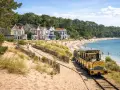 Petit train jaune longeant une plage de sable bordée de maisons colorées et d'une forêt.