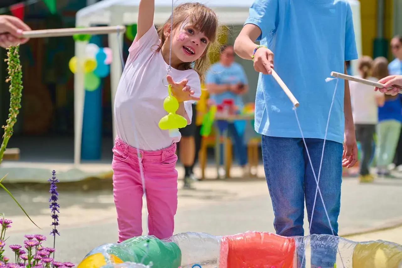 Une petite fille joyeuse participe à des jeux de kermesse pour tout petit, essayant d'attraper un jouet jaune avec une canne à pêche improvisée.