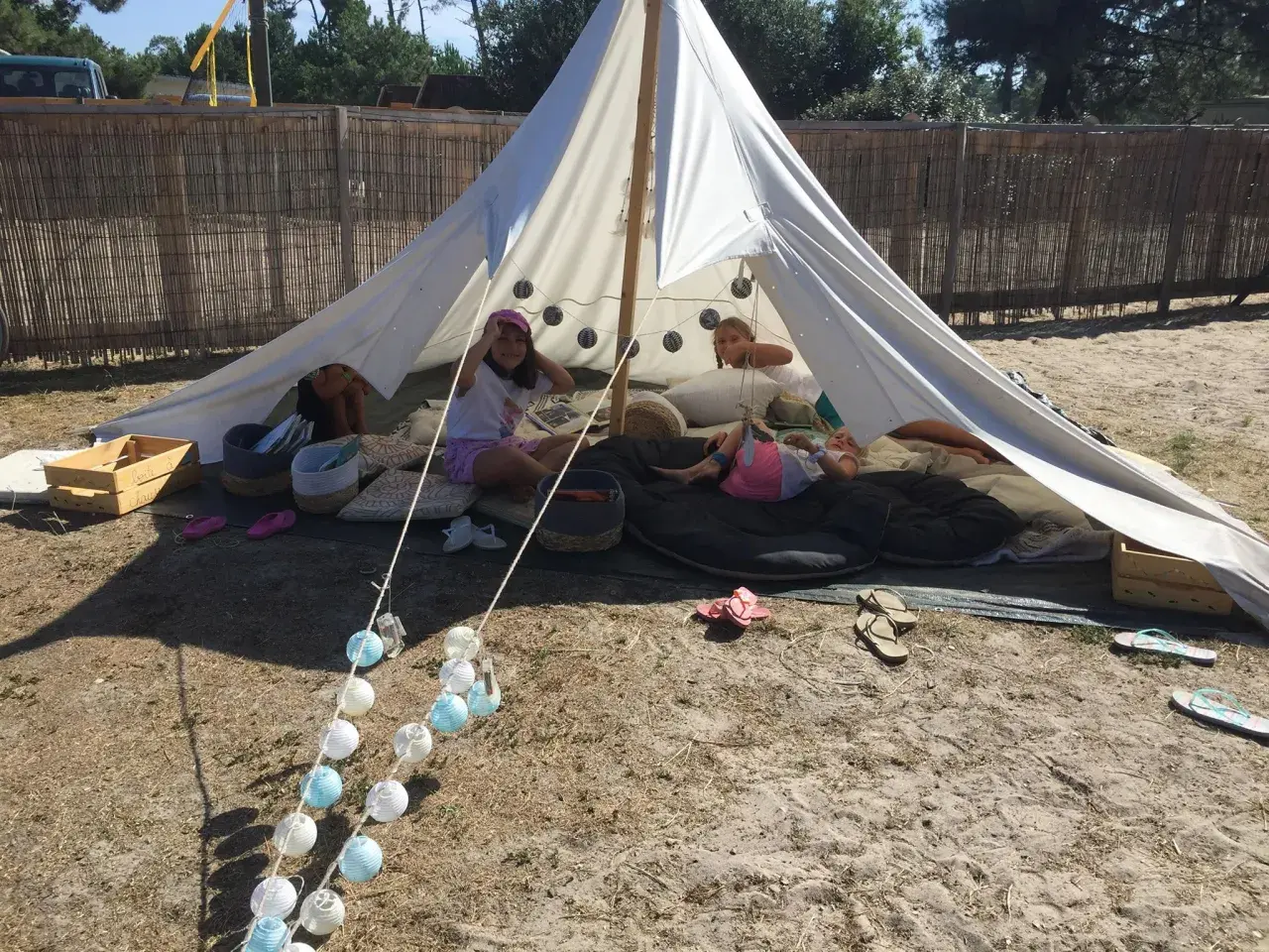 Enfants profitant d'une colonie de vacances camping dans une tente tipi, entourés de coussins et de guirlandes lumineuses.