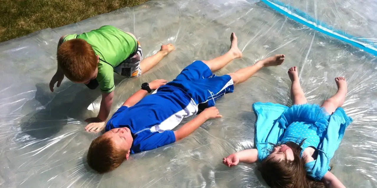 Trois enfants s'amusent sur un jeu d'eau pour kermesse, glissant sur une bâche mouillée sous le soleil.