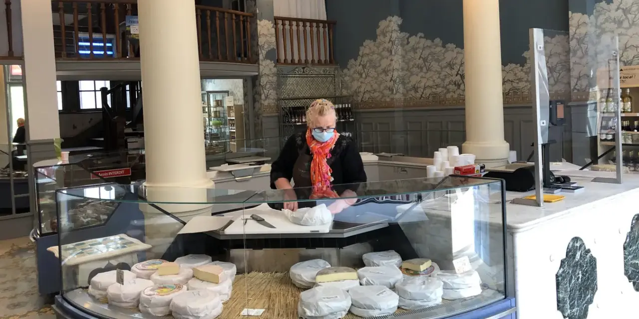 Une fromagère masquée prépare des fromages dans une vitrine réfrigérée.