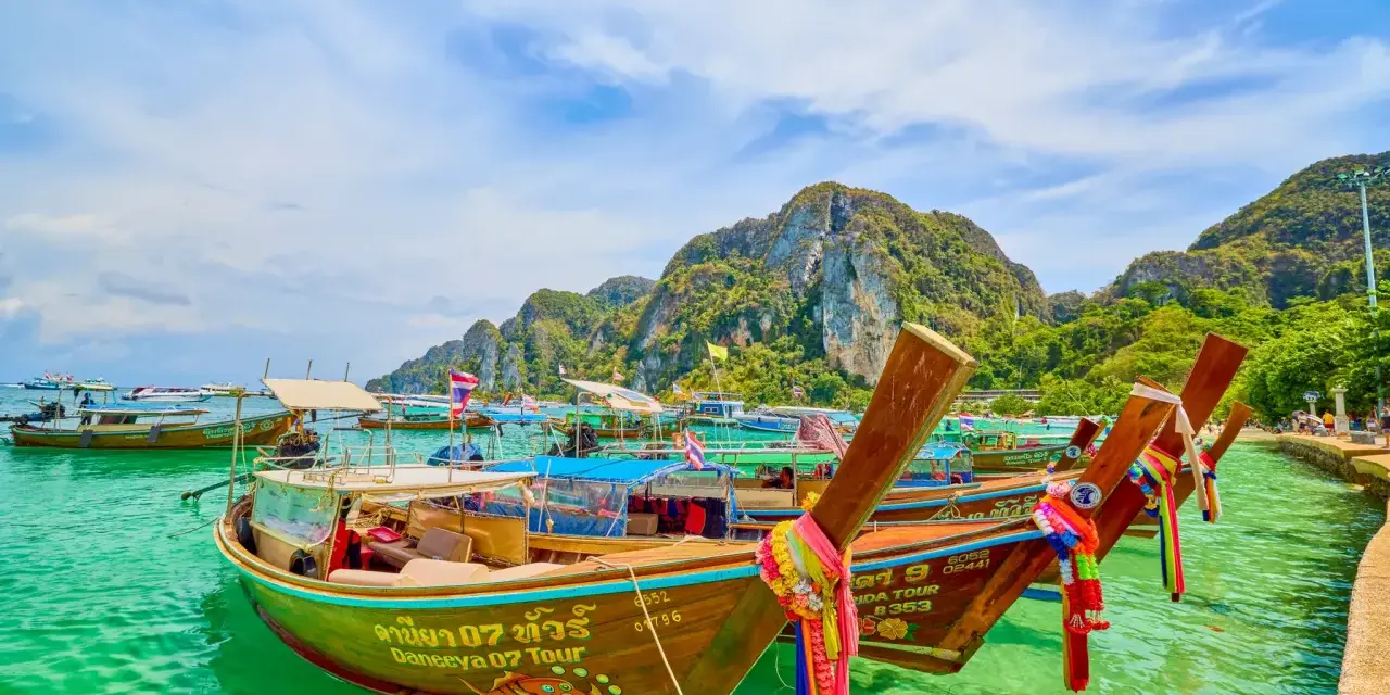 Bateaux traditionnels thaïlandais amarrés dans une eau turquoise, avec des montagnes verdoyantes en arrière-plan.