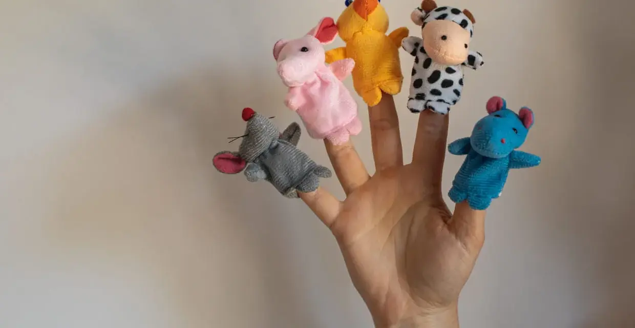 Main d'une personne avec cinq marionnettes à doigts en feutrine : souris grise, lapin rose, canard jaune, vache noire et blanche, hippocampe bleu.
