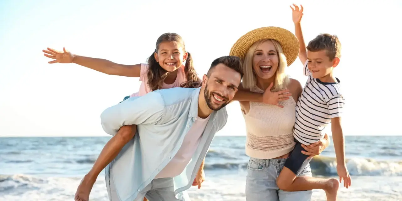 Famille souriante sur la plage, père portant sa fille sur le dos, mère tenant son fils.