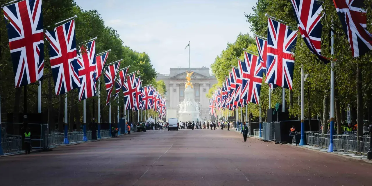 Allée bordée de drapeaux britanniques menant à un palais, avec des arbres et des gardes.