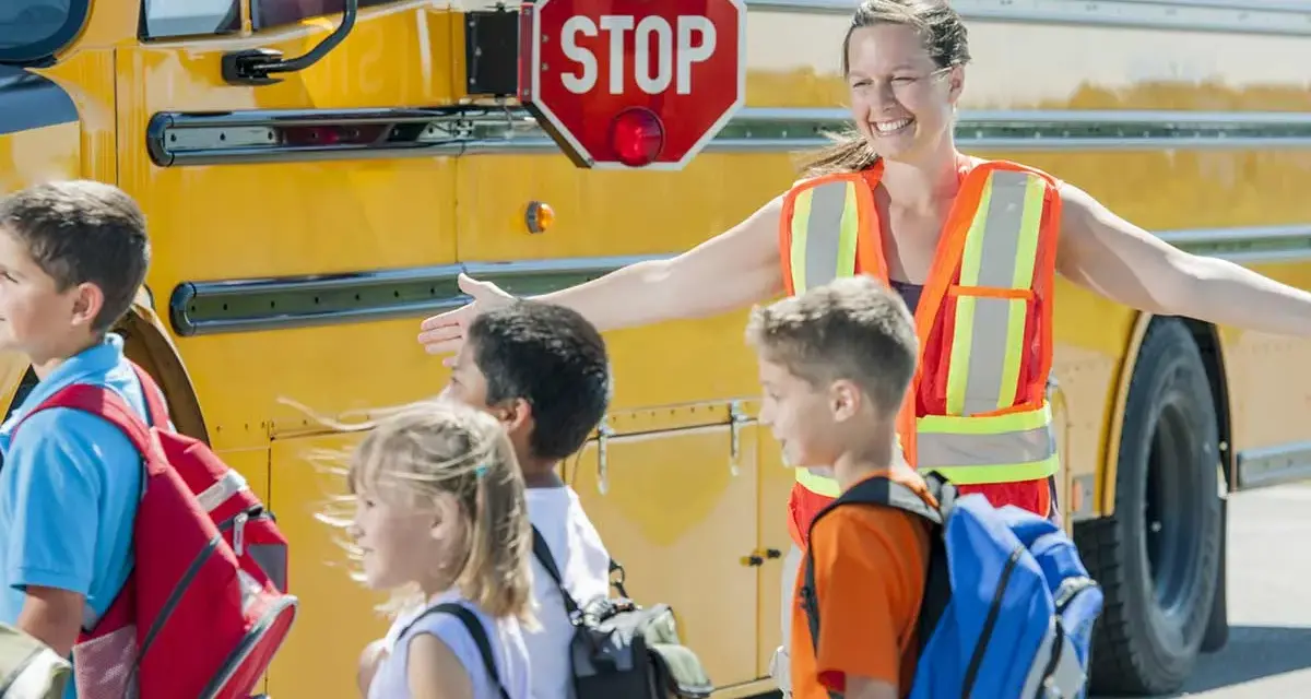 Des enfants montent dans un bus scolaire jaune, guidés par une adulte portant un gilet de sécurité orange.