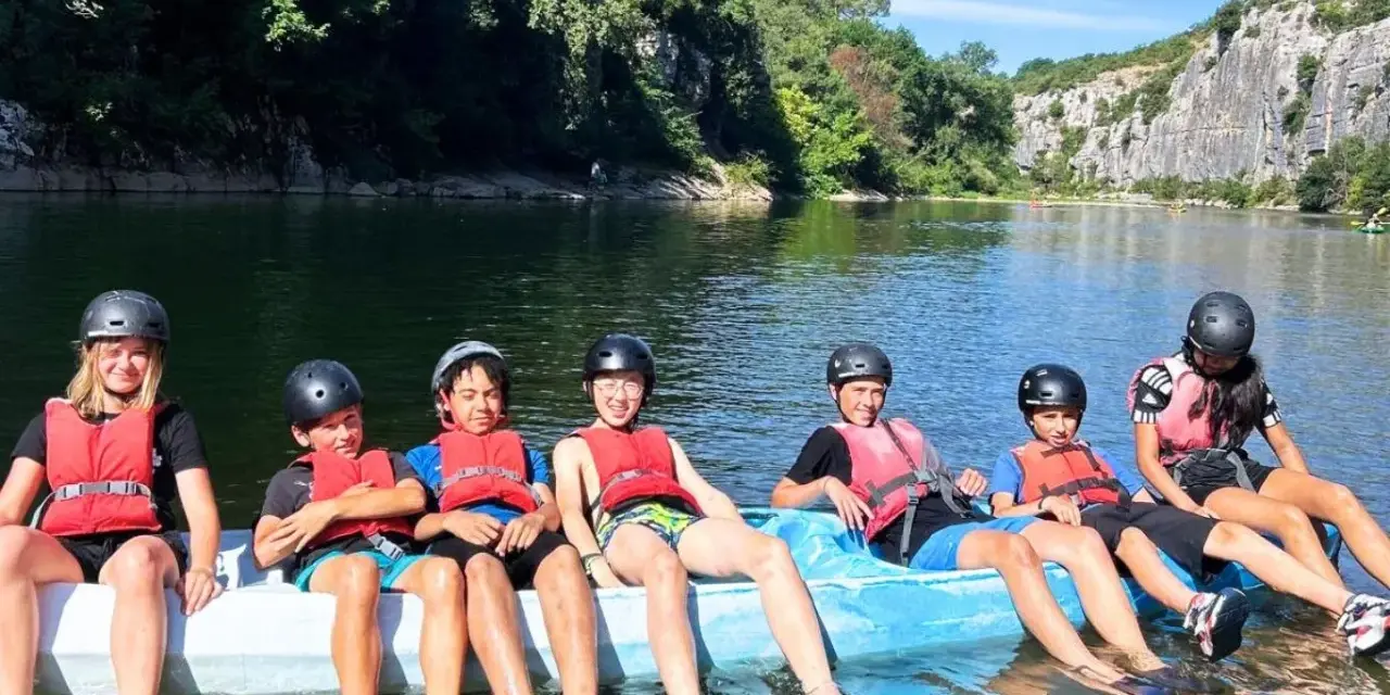 Des enfants en gilets de sauvetage et casques sur un raft bleu sur une rivière, entourés de verdure et de falaises.