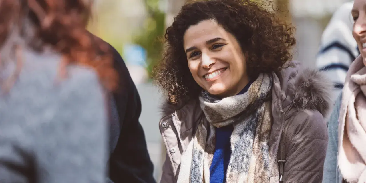 Une femme souriante aux cheveux bouclés porte une écharpe et une veste à capuche en fourrure.