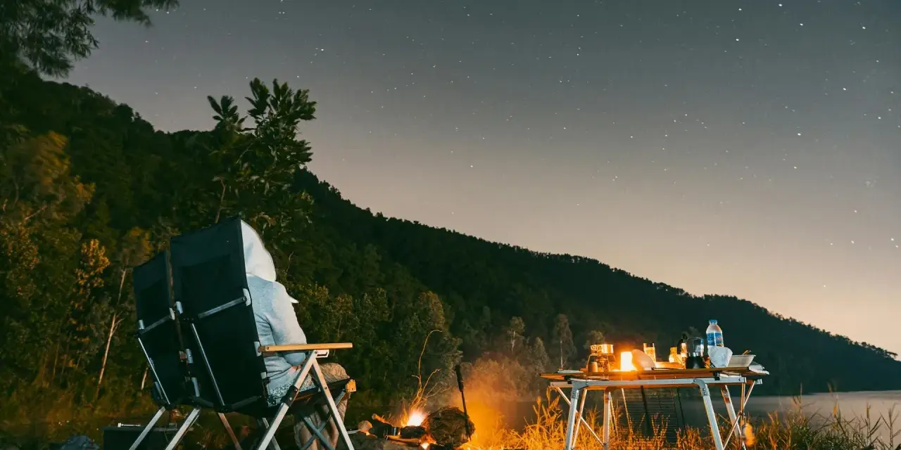 Personne assise près d'un feu de camp sous un ciel étoilé, avec une table de camping et un lac.