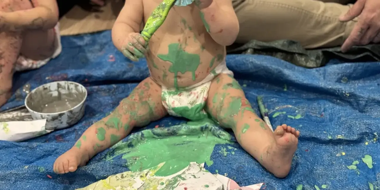 Bébé couvert de peinture verte, tenant un pinceau, assis sur une bâche bleue.