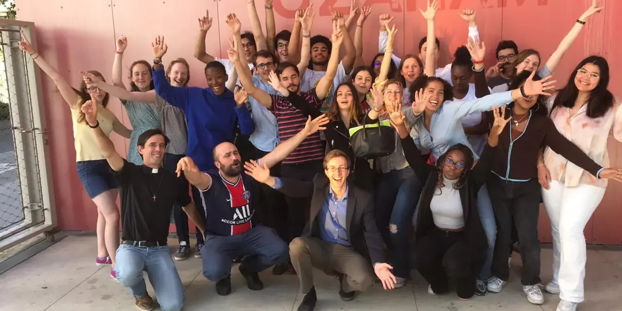 Un groupe de jeunes gens, dont un prêtre, pose joyeusement devant un mur rose avec le texte "MAISON OZANAM".