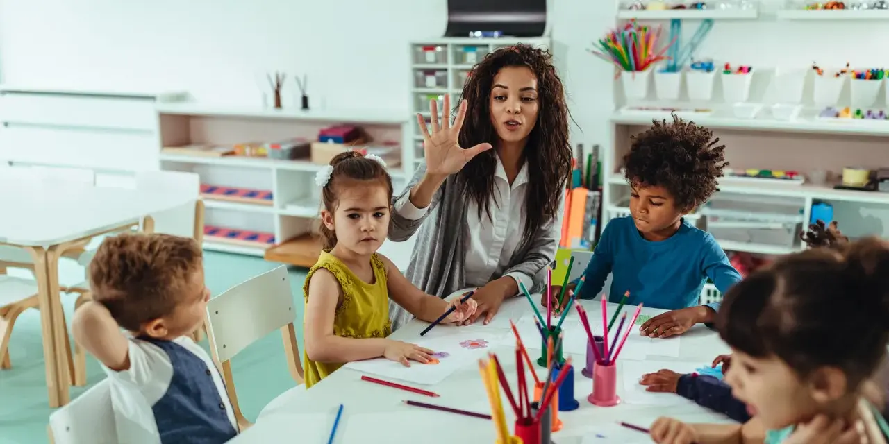 Une animatrice explique une activité créative à des enfants autour d'une table. Son rôle est d'éveiller leur curiosité.