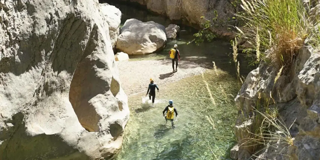 Trois personnes en combinaison font du canyoning dans une rivière aux eaux claires, entourées de falaises rocheuses.