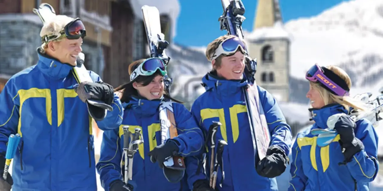 Quatre jeunes skieurs souriants en tenue bleue et jaune, portant des skis et des bâtons, dans une station de ski enneigée.