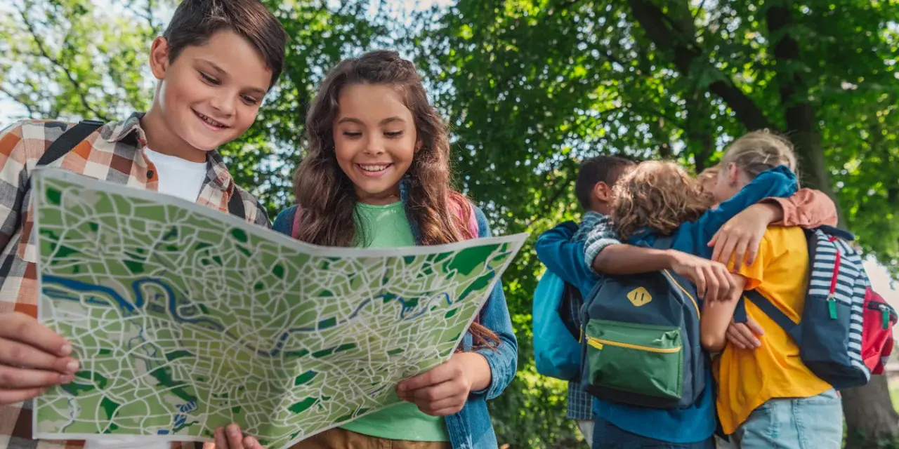 Deux enfants souriants regardent une carte dans un parc, d'autres enfants s'enlacent en arrière-plan.