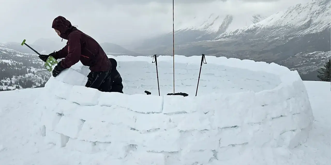 Person construisant un igloo en blocs de neige, avec des bâtons de ski plantés.