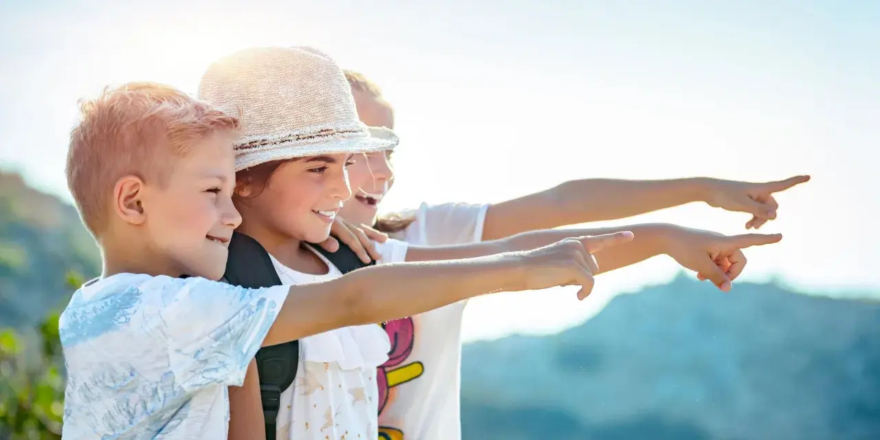 Trois enfants souriants, portant des sacs à dos, pointent du doigt vers la droite, sous un ciel ensoleillé.