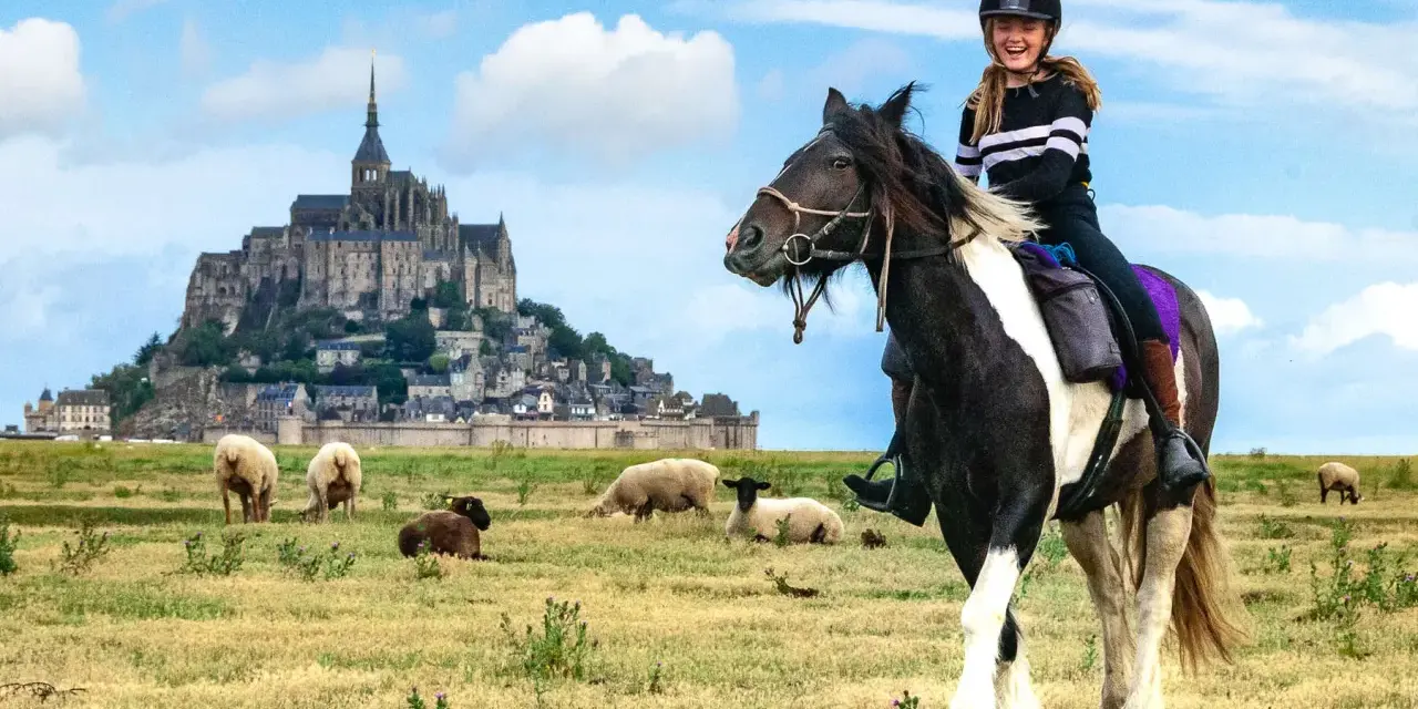 Une jeune fille rit à cheval devant le Mont-Saint-Michel, avec des moutons dans un pré.
