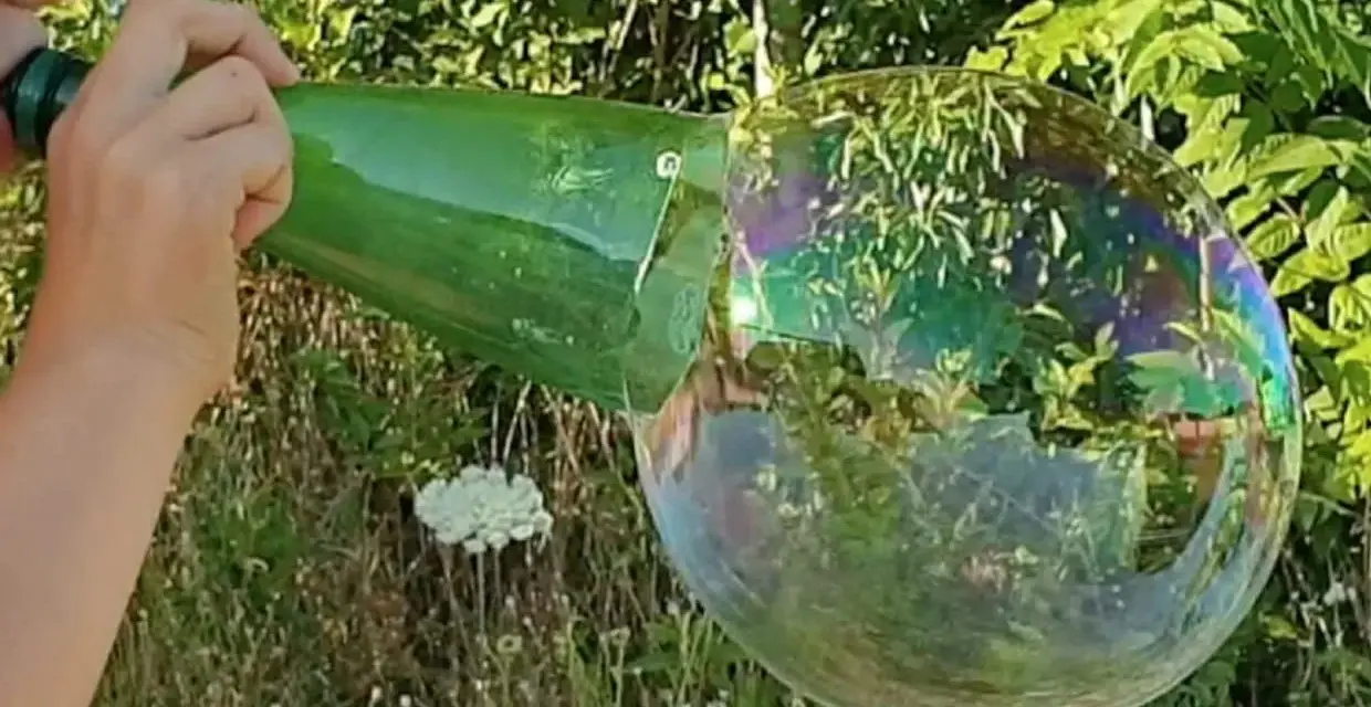 Main d'enfant soufflant dans une bouteille verte pour créer une bulle de savon géante irisée.