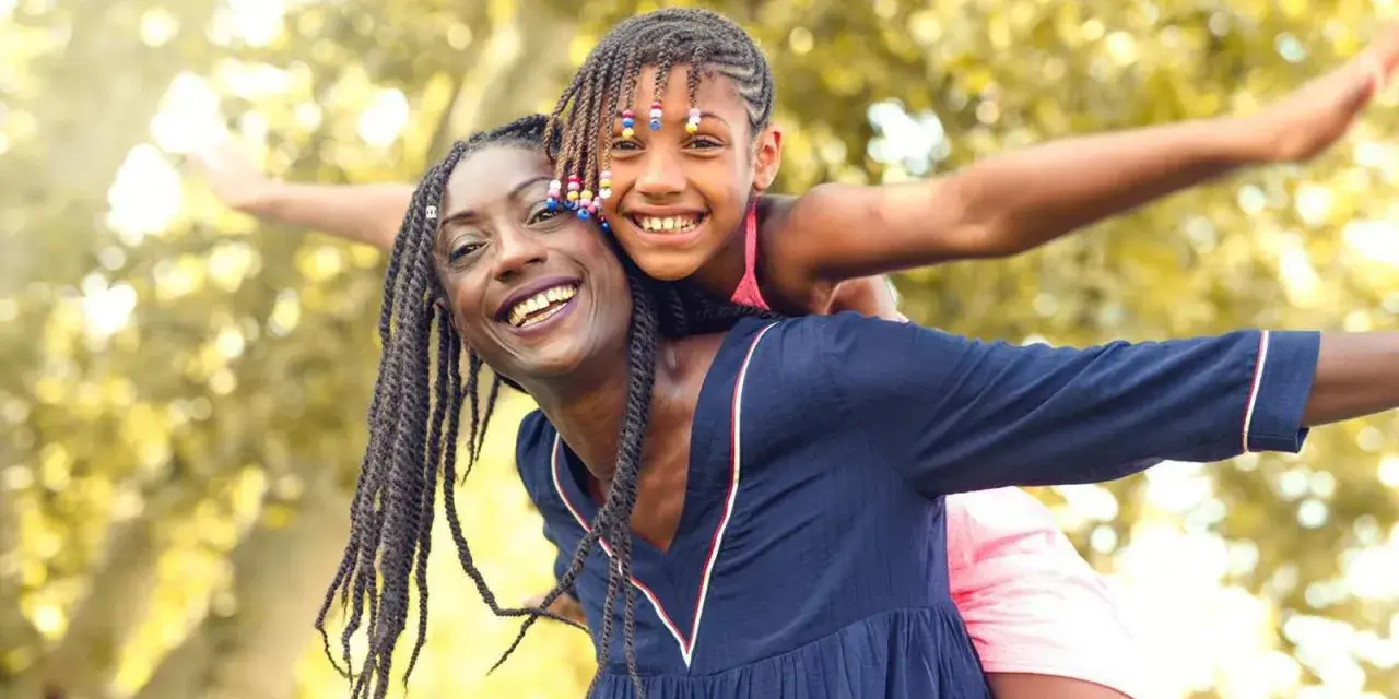 Une femme porte une enfant sur son dos, toutes deux souriantes, avec des arbres en arrière-plan.