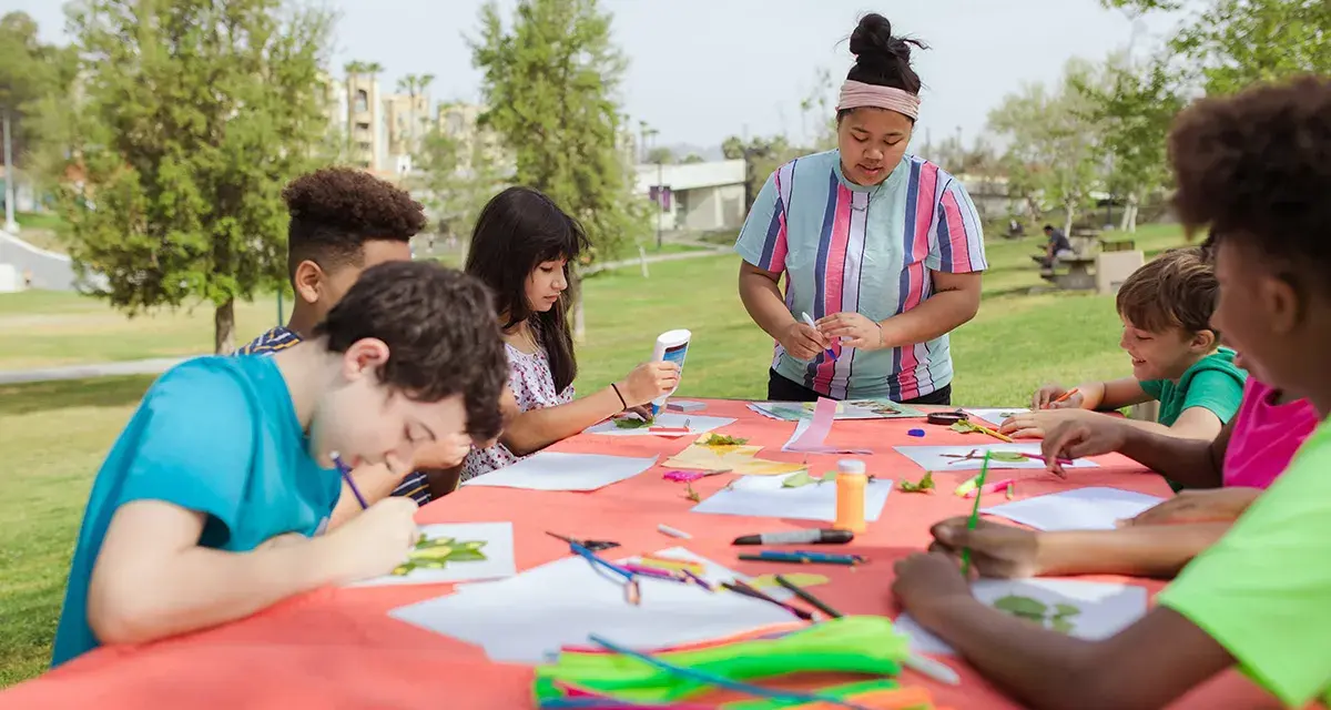 Des enfants créent des œuvres d'art en plein air autour d'une table recouverte d'une nappe orange.