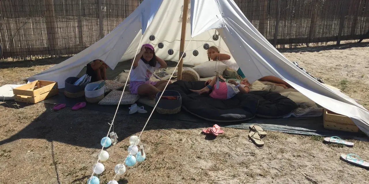 Enfants profitant d'une colonie de vacances camping dans une tente tipi, entourés de coussins et de guirlandes lumineuses.