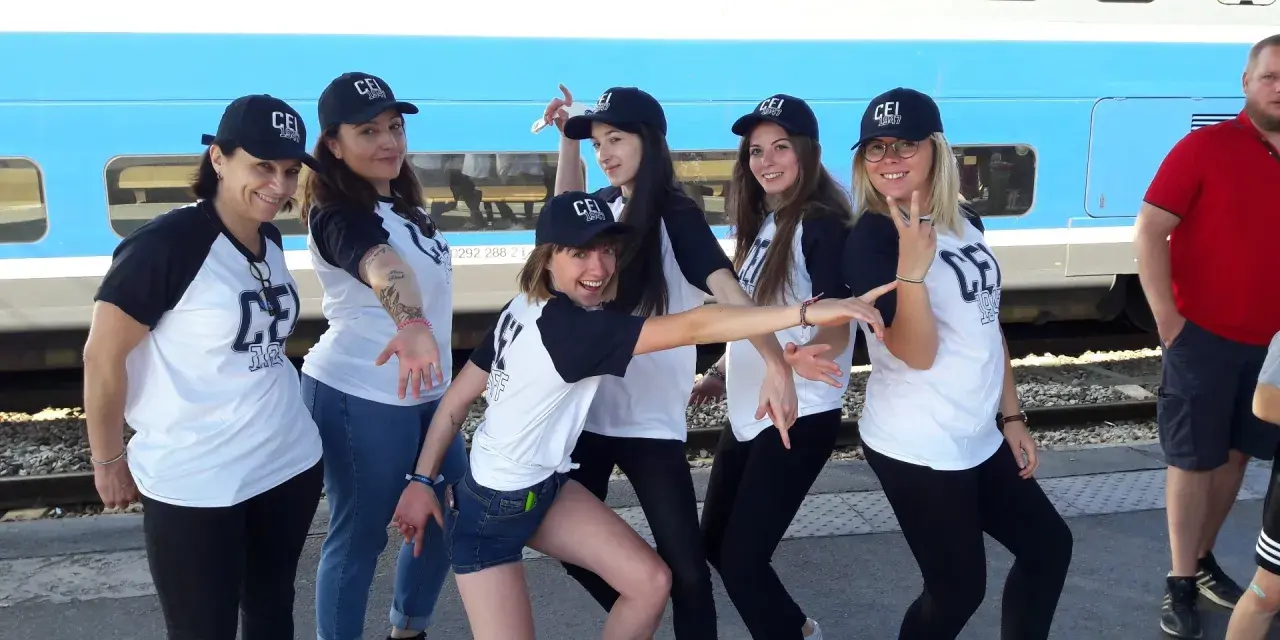 Groupe de jeunes femmes souriantes portant des casquettes et t-shirts "CEI 1947" devant un train.
